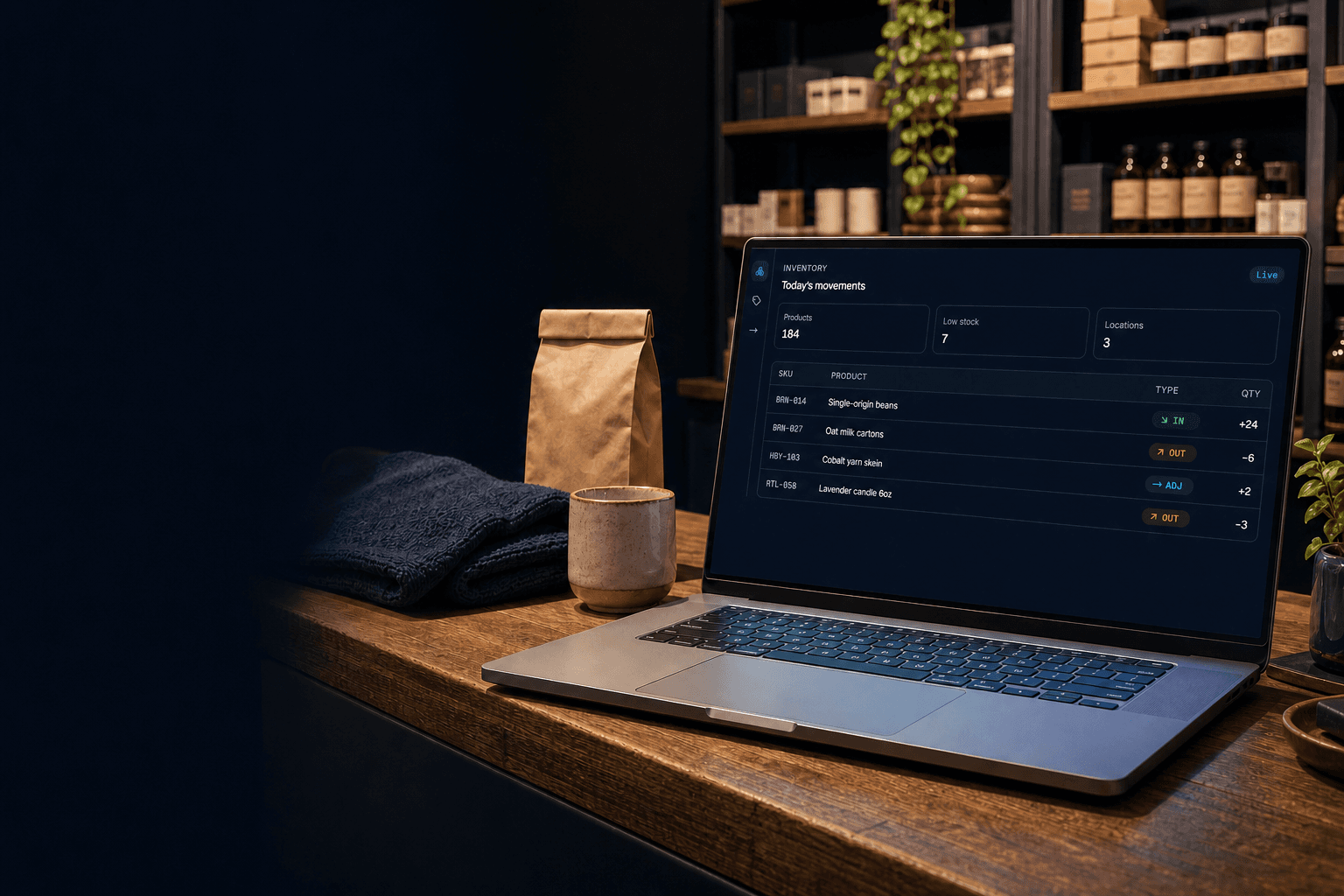 A laptop on a boutique shop counter showing the Digiventory inventory board with today's stock movements, beside a kraft-paper bag, ceramic mug, and folded textile.